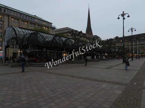 Place avec des arbres taillés et une structure en verre, à Hambourg, en Allemagne. Un clocher élancé se dresse en arrière-plan.