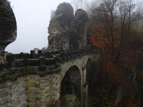 Pont de Bastei en Allemagne, entouré de rochers imposants et de feuillage automnal, sous un ciel brumeux.