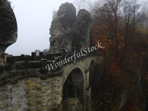 Pont de Bastei en Allemagne, entouré de rochers imposants et de feuillage automnal, sous un ciel brumeux.
