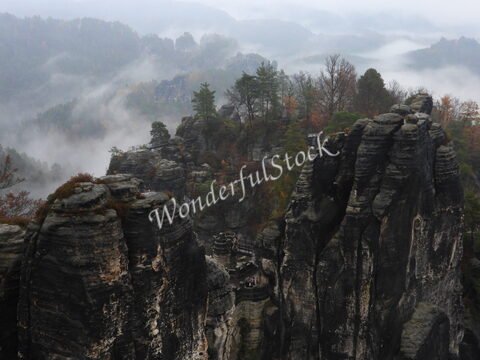 Rochers surplombant la brume dans la région de Bastei, en Allemagne, avec des arbres épars et une passerelle en bois visible.
