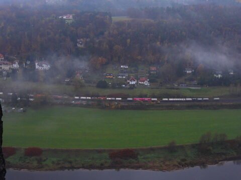 Un train circule le long d'un champ verdoyant près du pont Bastei en Allemagne, avec des maisons visibles en arrière-plan et de la brume.