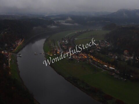 Vue sur le pont de Bastei en Allemagne, avec des maisons éclairées le long de la rivière et des collines environnantes sous un ciel nuageux.