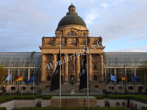 Bâtiment historique avec une grande coupole à Munich, Allemagne, entouré de drapeaux et d'une statue au premier plan.