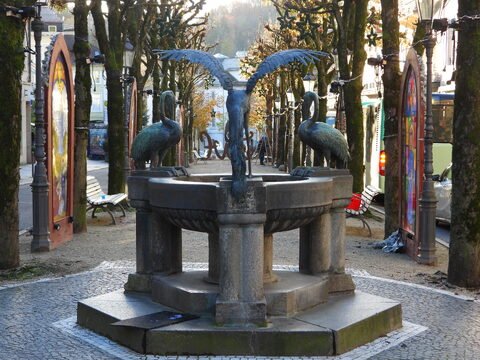Fontaine ornée de sculptures de cygnes à Baden-Baden, Allemagne, entourée d'arbres et de bancs, avec des affiches colorées en arrière-plan.