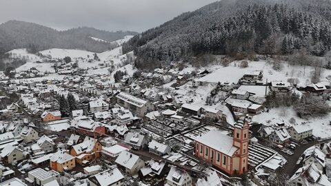Vue aérienne d'un village enneigé en Allemagne, avec des maisons aux toits blancs et une église à la façade rose.