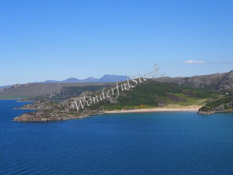 Plage bordée de rochers et de collines en Écosse. L'eau est calme et d'un bleu profond, sous un ciel dégagé.