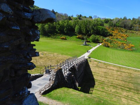 Un chemin menant à un monument dans un paysage verdoyant à Loch Ness, Écosse, avec des collines et des fleurs jaunes.
