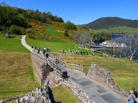 Un chemin pavé avec des visiteurs menant vers le Loch Ness en Écosse, entouré de collines verdoyantes et de fleurs jaunes.