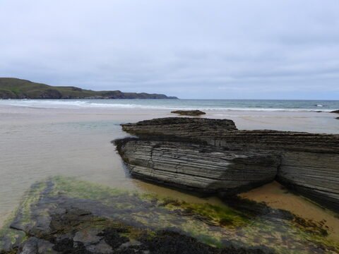Plage de Strathy en Écosse, avec des rochers striés et une mer calme sous un ciel nuageux.