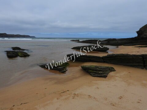 Plage de Strathy en Écosse, avec des rochers sur le sable et des vagues au loin sous un ciel nuageux.