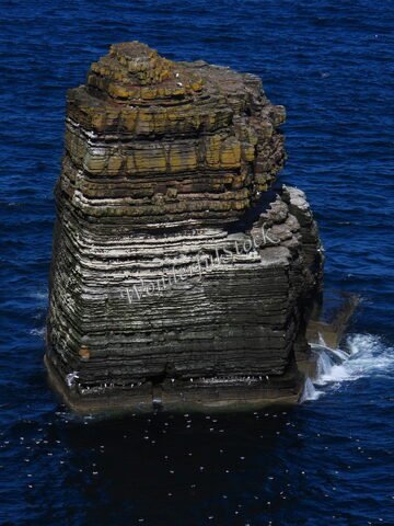 Un rocher strié émergeant de l'océan à Duncansby Head, Écosse, entouré d'eau bleue et de quelques oiseaux en vol.