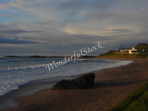 Plage de Ballycastle, Irlande, avec des vagues et des rochers au premier plan, et des maisons blanches en arrière-plan.
