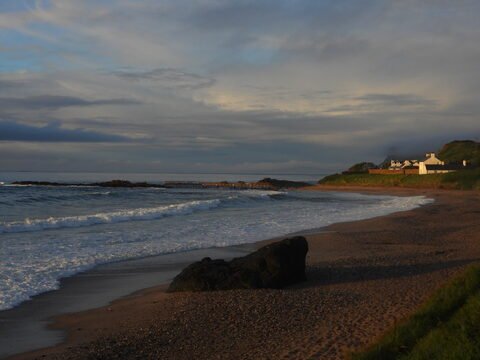 Plage de Ballycastle, Irlande, avec des vagues et des rochers au premier plan, et des maisons blanches en arrière-plan.