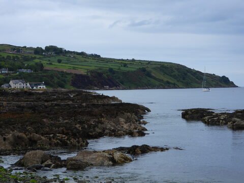 Bord de mer à Ballycastle, Irlande, avec des rochers et une colline verdoyante en arrière-plan, et un voilier sur l'eau.
