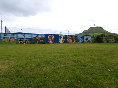 Mur peint avec des illustrations sportives et culturelles, avec une colline en arrière-plan, à Ballycastle, Irlande.