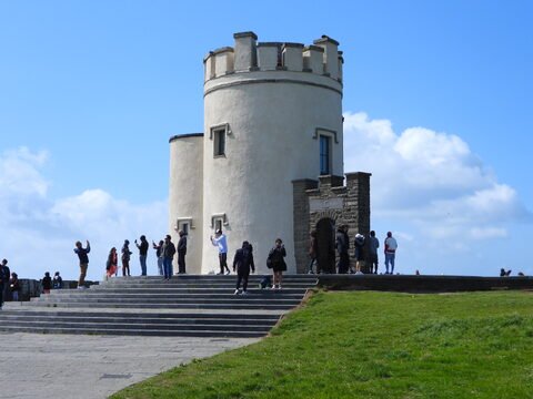 Un groupe de personnes se trouve près d'une tour sur les falaises de Moher, en Irlande. Le ciel est clair avec quelques nuages.
