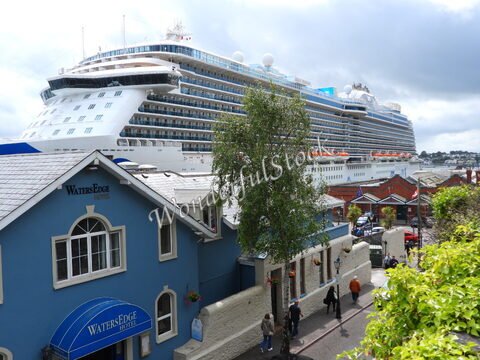 Un grand paquebot de croisière à Cobh, Irlande, avec des bâtiments colorés et des passants sur le trottoir.