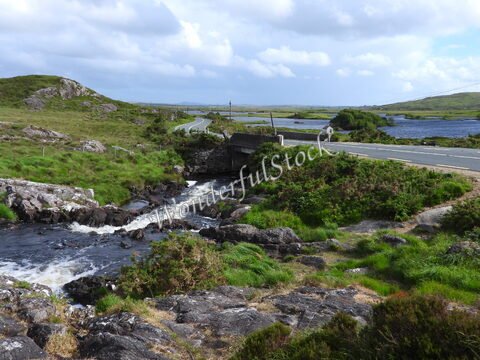 Un ruisseau traverse un paysage verdoyant avec des collines et une route à Connemara, Irlande.