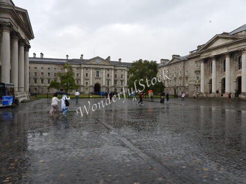Des personnes marchent sous la pluie dans une cour pavée à Dublin, Irlande, entourée de bâtiments historiques et d'arbres.