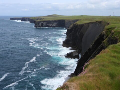 Falaises abruptes surplombant l'océan à Loop Head, Irlande, avec des vagues s'écrasant contre la roche et des prairies verdoyantes.