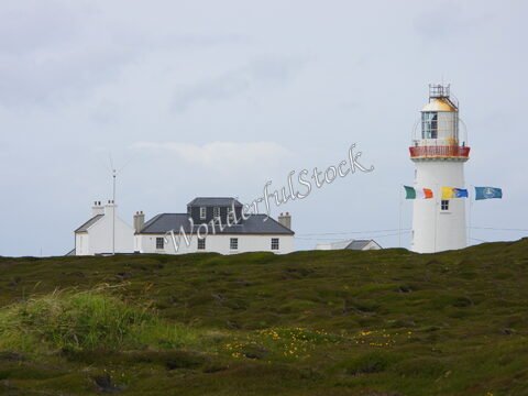 Phare sur Loop Head, Irlande, avec un bâtiment blanc et des drapeaux colorés, entouré de végétation.