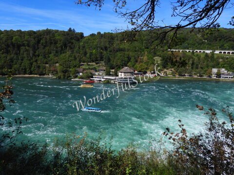 Chutes du Rhin en Suisse, avec des bateaux sur l'eau et des bâtiments en bordure de rivière, entourés de végétation.
