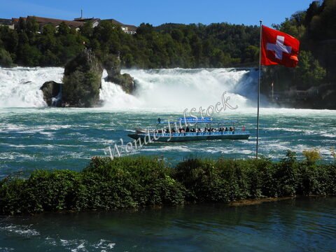 Bateau naviguant près des chutes du Rhin en Suisse, avec un drapeau suisse en premier plan et des arbres en arrière-plan.