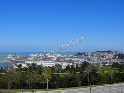 Vue sur le port d'Ancône en Italie, avec des navires et des grues, entouré d'une végétation verdoyante et d'une colline.