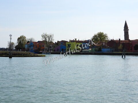 Des maisons colorées bordent l'eau à Murano, en Italie, avec un clocher visible en arrière-plan.