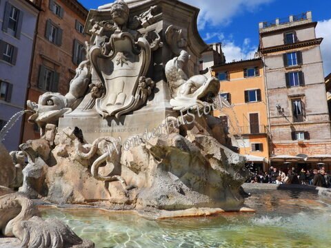 Fontaine ornée de sculptures à Rome, Italie, avec des éléments aquatiques et des bâtiments historiques en arrière-plan.