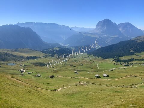 Paysage verdoyant à Seceda, Italie, avec des chalets, des collines et des montagnes en arrière-plan sous un ciel dégagé.