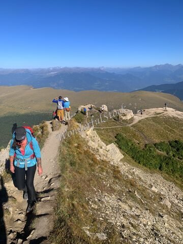 Randonnée sur un sentier étroit à Seceda, en Italie, avec des montagnes en arrière-plan et des personnes en train de marcher.