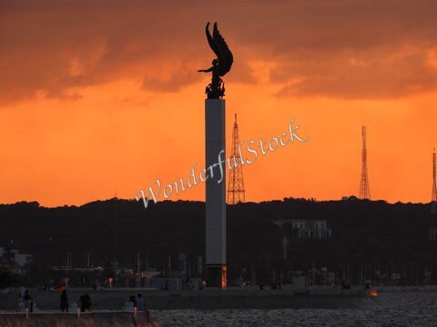 Monument avec ailes à Campeche, Mexique, silhouetté contre un ciel orange, entouré de collines et de structures en arrière-plan.