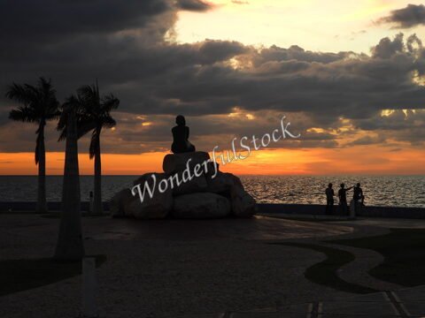 Silhouette d'une statue assise sur des rochers, avec des palmiers et un coucher de soleil à Campeche, Mexique.
