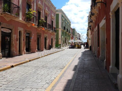 Rue pavée avec des bâtiments colorés à Campeche, Mexique, avec des parasols sur des tables et un ciel bleu partiellement nuageux.