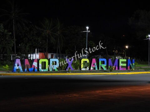 Lettrage coloré "AMOR X CARMEN" sur un rond-point à Carmen, Mexique, avec des palmiers en arrière-plan sous un ciel nocturne.