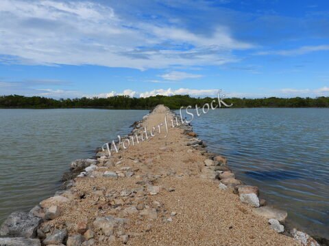 Un chemin de pierres et de sable s'étend sur l'eau à Santa Clara, au Mexique, entouré de végétation et sous un ciel partiellement nuageux.