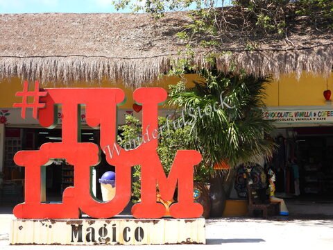 Tulum au Mexique avec un grand panneau rouge "#TULUM Magico" devant un magasin, entouré de verdure et d'une toiture en chaume.