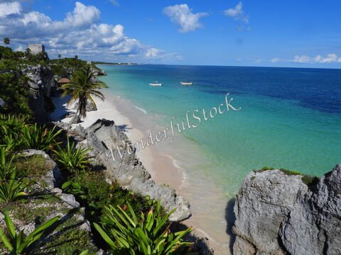 Plage de sable avec des rochers et des palmiers à Tulum, Mexique, bordée par une mer turquoise et un ciel bleu parsemé de nuages.