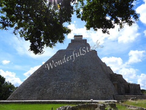 Pyramide d'Uxmal, Mexique, avec un ciel bleu parsemé de nuages et des arbres en ombre au premier plan.