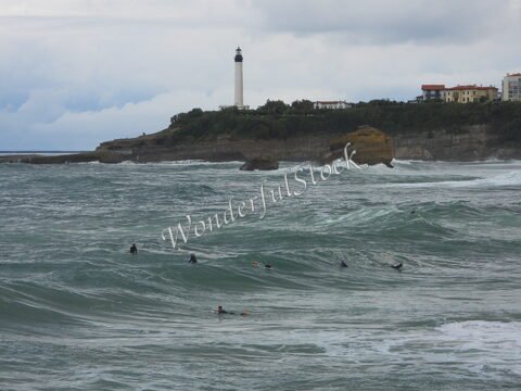 Des surfeurs dans les vagues à Biarritz, France, avec un phare en arrière-plan et des falaises au loin.