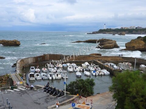 Bateaux amarrés dans un port à Biarritz, France, avec des rochers et un phare en arrière-plan, sous un ciel nuageux.