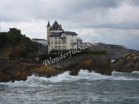 Maison surplombant la mer à Biarritz, France, avec des vagues déferlant contre les rochers et un ciel nuageux en arrière-plan.