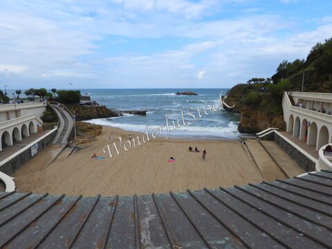 Plage de Biarritz, France, avec des personnes sur le sable et des vagues au loin, entourée de structures en béton et de verdure.
