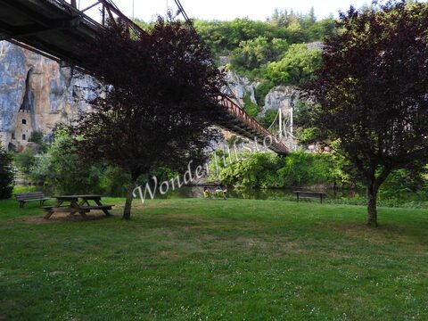 Pont suspendu au-dessus d'un terrain verdoyant à Bouzies, France, avec des arbres et une rivière à proximité.