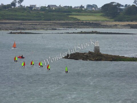 Des voiliers aux voiles vertes et rouges naviguent près d'un phare sur la côte à Aber Wrach, en Bretagne, France.