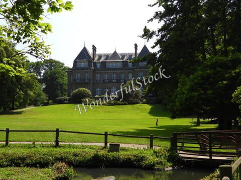 Un bâtiment en pierre se dresse sur une pelouse verte à Dinard, France, avec des arbres autour et un petit pont en bois à proximité.