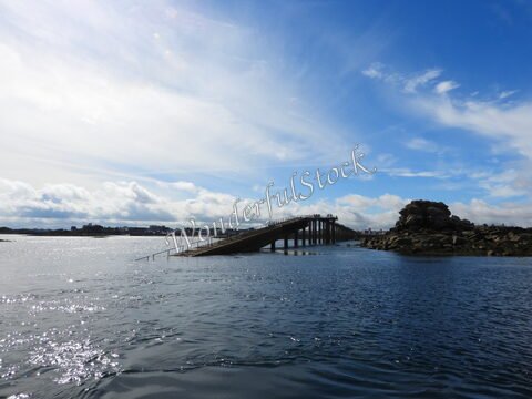 Un pont en bois s'étend sur l'eau à l'île de Batz, en Bretagne, France, avec des rochers visibles et un ciel nuageux.