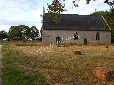 Église en pierre avec fenêtres gothiques à Locmariaquer, Bretagne, entourée de champs de foin et de quelques arbres.