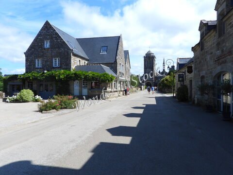 Rue pavée avec des maisons en pierre et une église à Locronan, France. Des fleurs bordent le chemin sous un ciel partiellement nuageux.